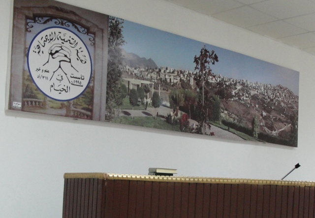 شعارهيئة التنمية الأجتماعية في الخيام إلى جانب صورة كبيرة لبلدة الخيام